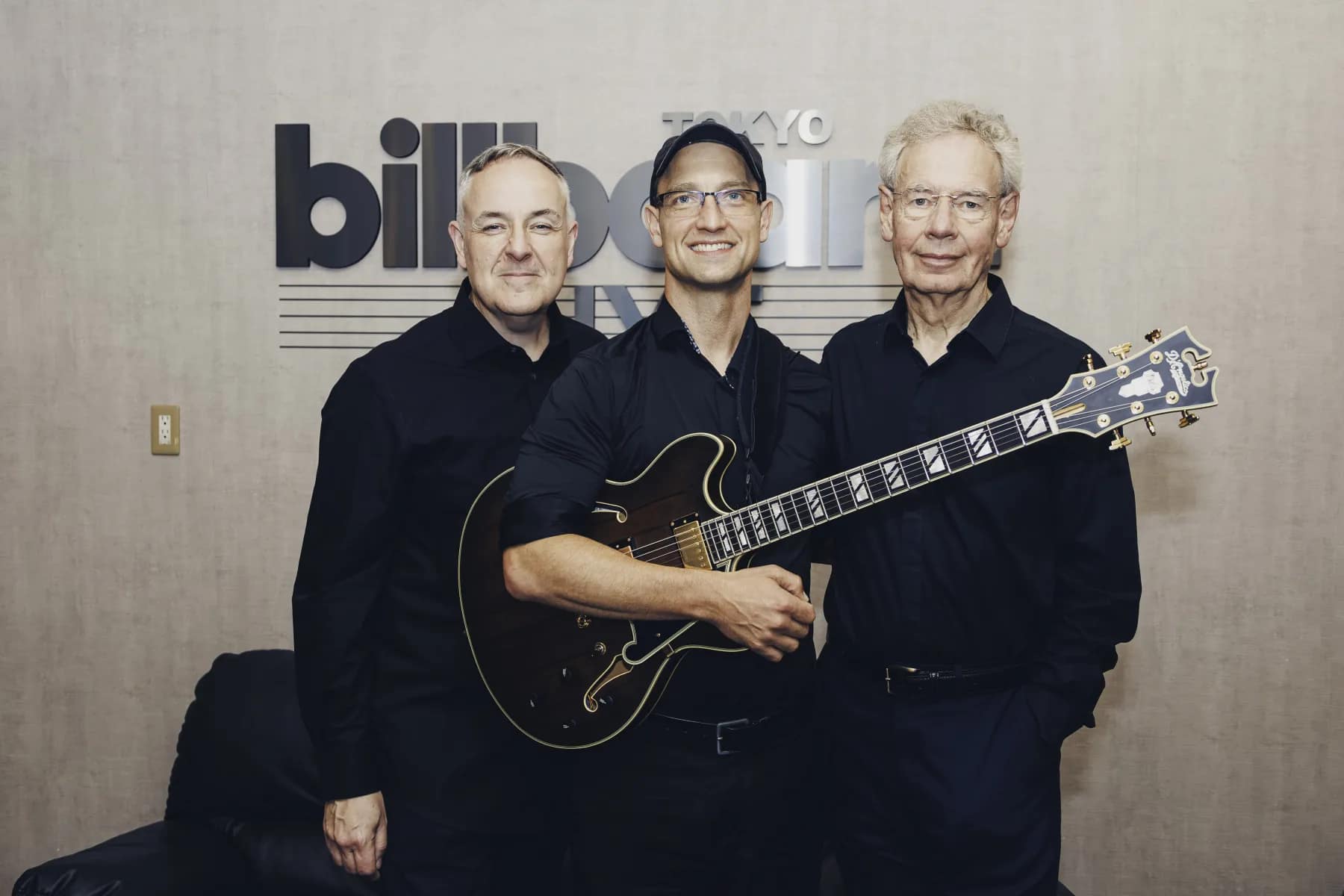 Pete Roth Trio standing together with guitar in front of the Billboard Live Tokyo wall.