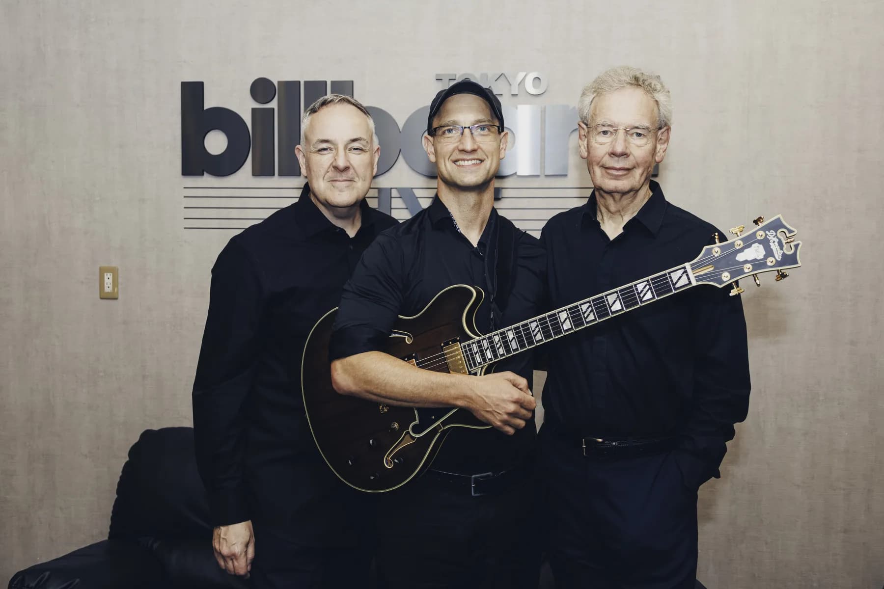 Pete Roth Trio standing together with guitar in front of the Billboard Live Tokyo wall.
