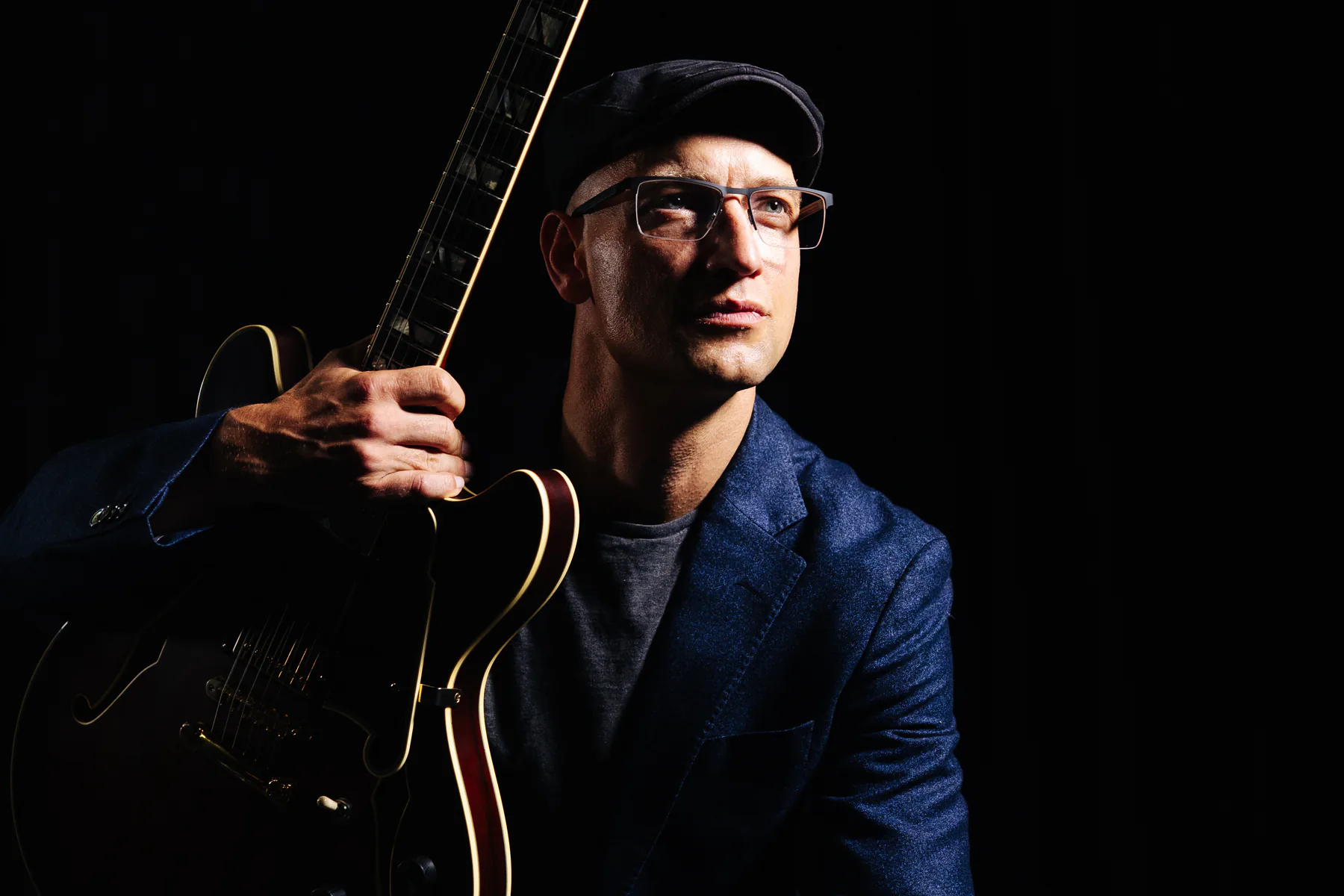 Pete Roth holding his guitar in a close colour portrait against black.